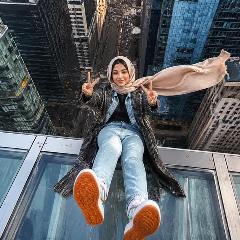 一张超写实都市高空摄影，纵向构图，广角镜头，俯拍视角，一位年轻中东/南亚气质女性坐在摩天大楼玻璃天窗的金属边缘上，双腿自然向前伸出悬空，鞋底朝向镜头形成强烈透视 - AI生成图片 1