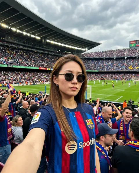 一张高真实感、社交媒体自拍风格的年轻女性半身近景照片，拍摄于一座座无虚席的大型足球场看台内，背景是绿茵球场、密集观众席与弧形球场顶棚，现场比赛日氛围浓厚。主角为 - AI生成图片 1