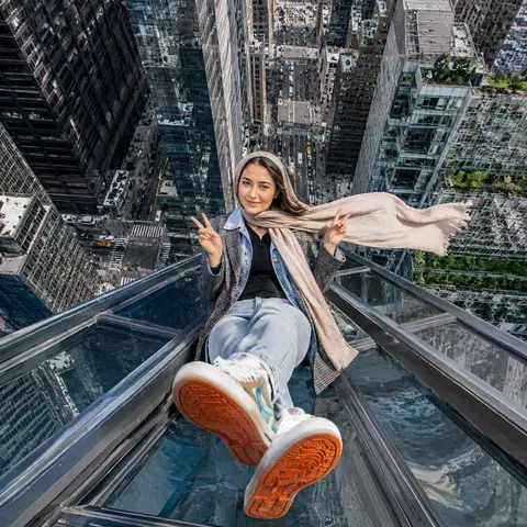 一张超写实都市高空摄影，纵向构图，广角镜头，俯拍视角，一位年轻中东/南亚气质女性坐在摩天大楼玻璃天窗的金属边缘上，双腿自然向前伸出悬空，鞋底朝向镜头形成强烈透视 - AI生成图片 2